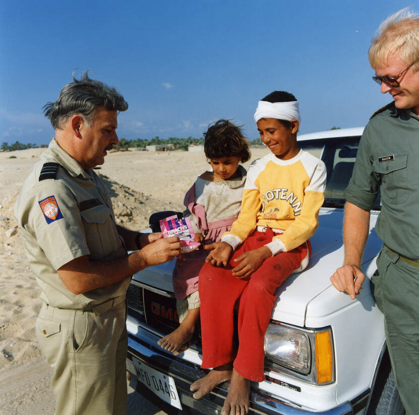 Kleurenfoto, buiten in de woestijn. Twee kinderen zitten op de motorkap van een witte jeep, één van hen heeft een verband om zijn hoofd. Zij lachen en kijken naar het snoep dat de militair links aan hen geeft. Deze militair draagt een beige uniform met op zijn rechtermouw het oranje-blauwe embleem van de operatie. Rechts naast de motorkap kijkt een blonde militair glimlachend toe. Op de achtergrond een woestijnlandschap met in de verte een oase.