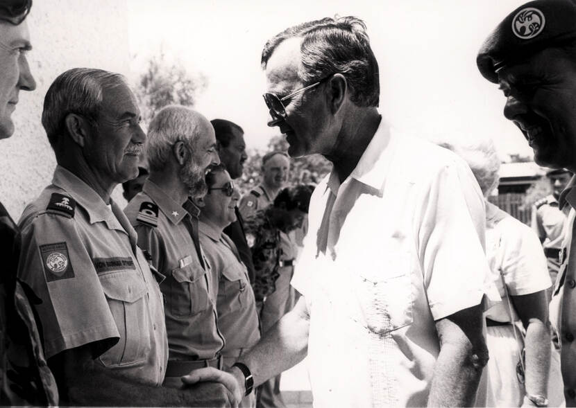 Zwart-witfoto. Links staan hogere militairen in een rij opgesteld. De tweede van links heeft een snor en schudt de hand van de man die voor hem staat. Deze draagt een wit overhemd met korte mouwen en een zonnebril. De andere militairen verderop in de rij lachen om iets dat zich achter de man in het wit afspeelt.