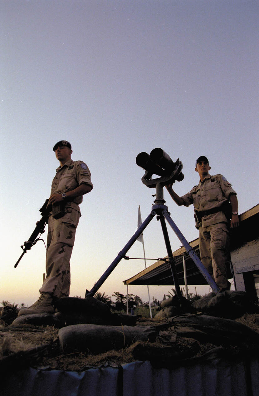 Kleurenfoto, buiten en van onderaf genomen. Twee militairen in beige uniformen staren voor zich uit. De linker heeft een geweer vast met de loop naar beneden, de rechter houdt zijn rechterhand op een grote verrekijker. De verrekijker staat op een driepoot. Op de achtergrond een gebouw met uitlopende overkapping en de toppen van een paar bomen.