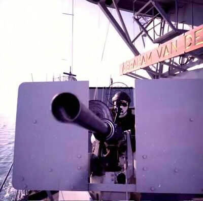 Kleurenfoto, buiten aan boord van een marineschip. Iemand zit achter een grote mitrailleur en richt de loop tussen twee metalen platen door richting de camera. De persoon draagt een helm en een witte bivakmuts. Voor zijn gezicht is een groot, rond vizier. Boven hem een houten bord met de naam van het schip, te lezen is: Abraham van de.