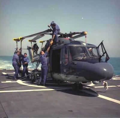 Kleurenfoto, buiten op het dek van een marineschip. Een helikopter staat geparkeerd op het dek, het is met een paar kabels vastgemaakt. De wieken zijn richting de staart ingeklapt, twee mannen zetten deze met stangen vast. Een man staat bovenop de helikopter en reikt iets naar een vierde man die naast het toestel staat. Alle mannen dragen blauwe overalls.