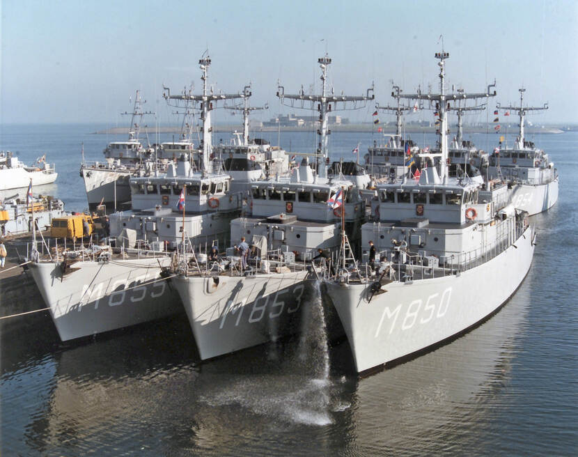 Kleurenfoto, buiten in een haven. Drie grijze marineschepen liggen tegen elkaar aan, op de dekken staan enkele personen, vanaf het rechter schip valt water de haven in. Op de zijkanten aan de voorkant van de schepen staan van links naar rechts M855, M853 en M850 geschilderd. Achter de schepen nog een rij van drie, waarvan de meest rechter M854 op de zijkant geschilderd heeft. Hier en daar wapperen kleine vlaggetjes.