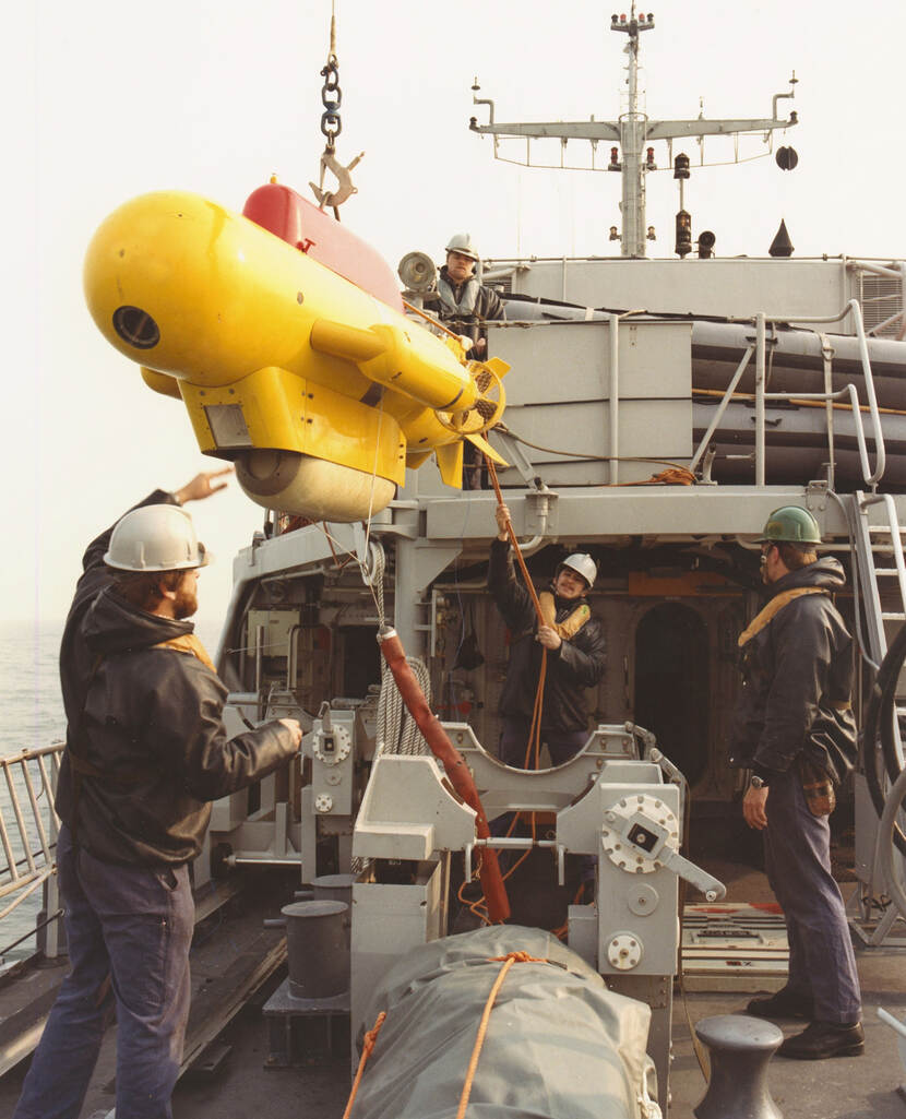 Kleurenfoto, op het dek van een marineschip. Drie mannen staan op het dek, de achterste houdt een touw vast, de voorste reikt met zijn linkerhand naar boven. Boven hen hangt een kleine gele onderzeeër aan een haak, aan de onderkant zit het vast aan een kabel. Een vierde man staat op een verhoging achter de onderzeeër. Alle vier de mannen dragen een helm, een kleine zwemvest en plastic regenjassen.