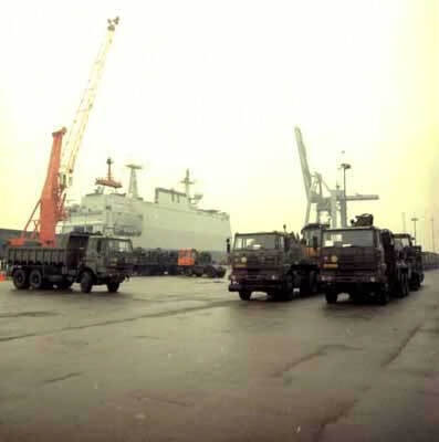 Kleurenfoto, buiten op een kade. Drie legervrachtwagen staan op een kade. Links in de achtergrond is een marineschip te zien, naast een hoge oranje hijskraan. Rechts in de achtergrond een hogere grijze hijskraan.