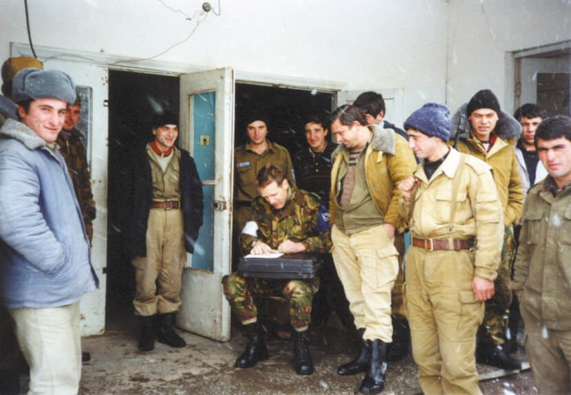 Kleurenfoto, buiten bij de ingang van een gebouw. Op een stoel zit een militair in camouflagekleding, hij heeft een witte band om zijn rechtermouw. Hij heeft een koffer op zijn schoot, die hij als tafel gebruikt om iets te schrijven. Om hem heen staan twaalf mannen, allemaal in dikke jassen, sommigen met een grote bontmuts op. Sommigen kijken in de camera, anderen naar de schrijvende militair. Het sneeuwt.
