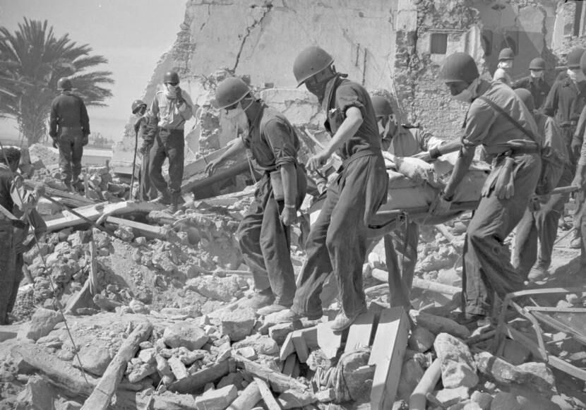 Zwart-witfoto, vijf militairen lopen over puin van ingestorte gebouwen en dragen een brancard. Om hen heen zijn andere militairen en burgers in het puin aan het zoeken.