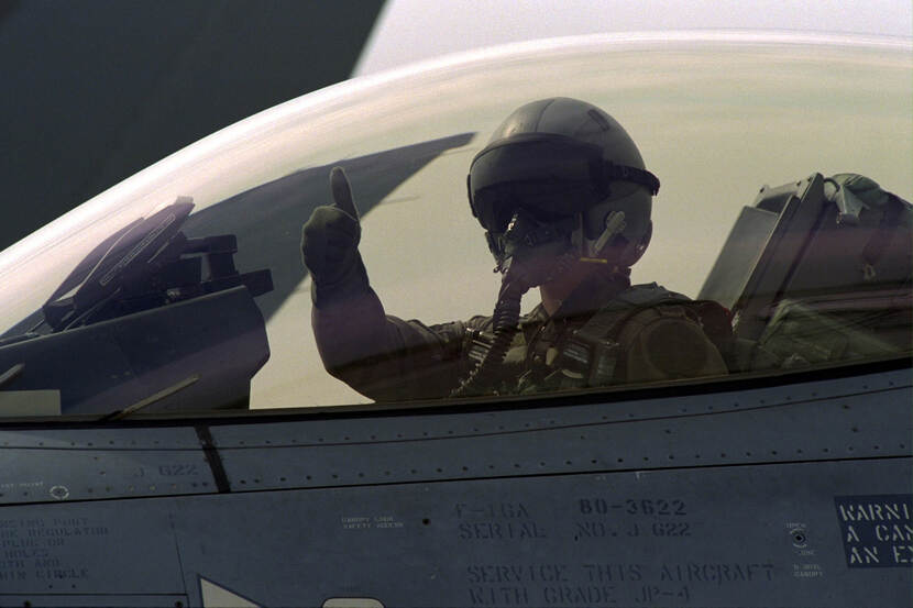 Kleurenfoto, buiten op een vliegveld. Een militaire piloot zit in de cabine van een militair vliegtuig, met gesloten koepel. Hij draagt een helm met donker vizier en een zuurstofmasker, hij steekt zijn rechterduim omhoog.