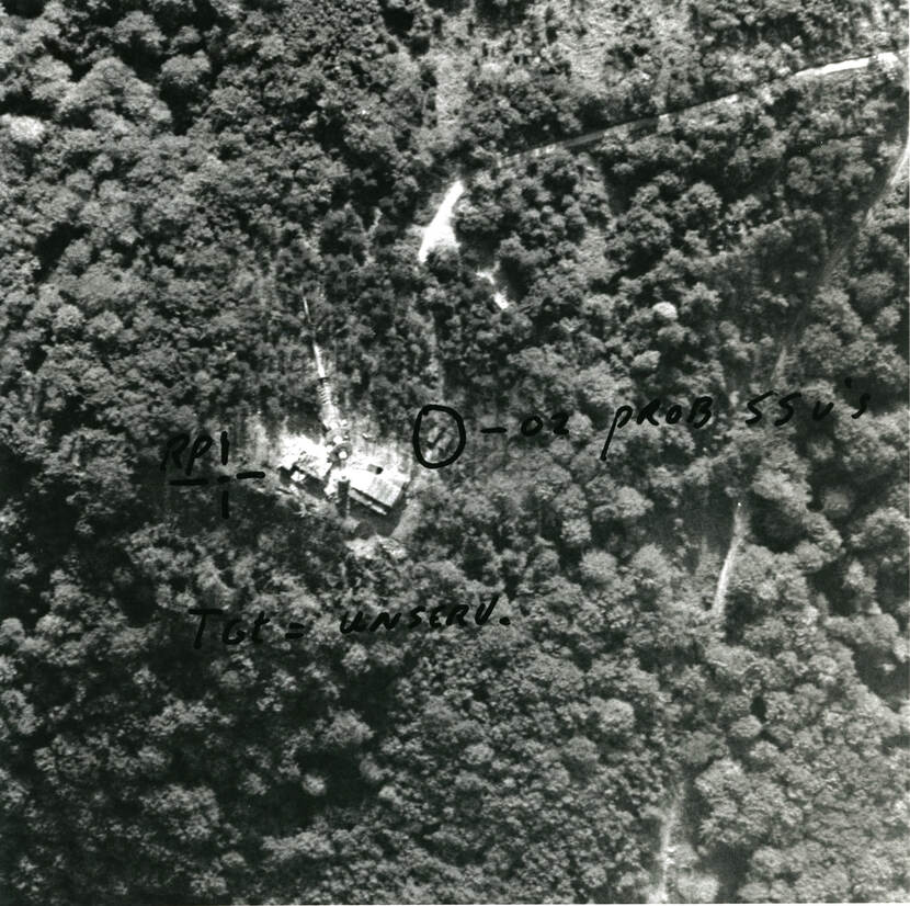 Zwart-witfoto, buiten in een bos. Luchtfoto van bovenaf genomen. Op de grond tussen de bomen ligt een communicatiemast naast de installatie waar het bovenop hoort. Daarnaast ligt iets dat door iemand op de foto met stift is omcirkeld en van onduidelijk commentaar is voorzien.