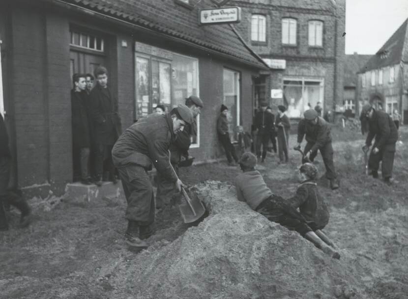Zwart-witfoto, vier militairen scheppen zand in een opengebroken straat. Op de zandhoop zitten twee kinderen, andere kinderen staan eromheen en kijken toe.