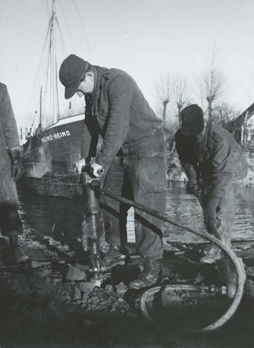 Zwart-witfoto, een militair staat op een kade langs het water, hij hanteert een drilboor. Rechts van hem een militair die aarde schept, links iemand die toekijkt. Op de achtergrond in het water een schip met de naam Heinz Heino.