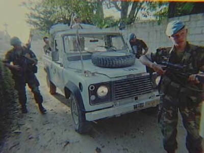 Kleurenfoto, buiten op straat. Vier militairen in groene camouflagekleding staan voor, naast en achter een witte jeep die onder het stof zit. Alle militairen dragen een blauwe baret. De voorste militair houdt een geweer in de aanslag, de militair links houdt een geweer omhoog en stapt naar voren. Op de achtergrond een muur en bomen, op de motorkap van de jeep ligt een reservewiel.