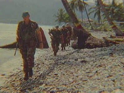 Kleurenfoto, buiten op een strand. Een groepje van vier militairen in groene camouflagekleding loopt richting de camera, alle militairen dragen een wapen over de schouder. Links van hen is het water, rechts is een kiezelstrand zijn palmbomen, sommigen hebben een rode en een gele verfstreep op de bast. Op de achtergrond meer palmbomen.