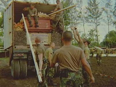 Kleurenfoto, buiten. Vijf militairen in groene camouflagekleding staan achter een open vrachtwagen. De vrachtwagen ligt vol met houten palen, latten en kisten, twee militairen zitten en staan op de lading en laden deze spullen uit. Eén militair reikt naar een uitgestoken paal uit, een ander heeft er één vast. Op de achtergrond lopen drie militairen met dezelfde spullen en zijn hoge bomen te zien.