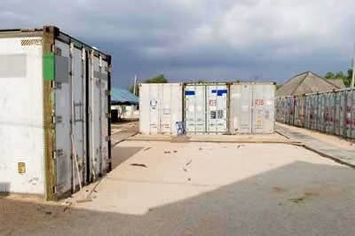 Kleurenfoto, buiten. Links een witte container, in het midden drie soortgelijke containers, rechts een lange rij containers. In de achtergrond, half verscholen achter de containers, een paar huizen. De lucht daarboven is bedekt met donkere wolken.