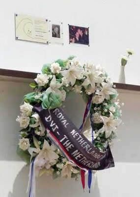 Kleurenfoto. Een krans van witte bloemen hangt aan een muur, er hangt een zwart lint overheen. Daarboven aan de muur hangen twee kleine foto’s en een plaquette. Rechts daarvan op een richel een vaas met één bloem.