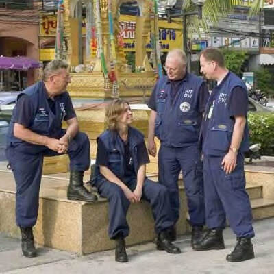 Kleurenfoto. Drie mannen en een vrouw zijn buiten op straat, de vrouw zit op een verhoging, de mannen staan om haar heen, zij dragen allemaal blauwe kleding. De linker man heeft zijn linkvoet op de verhoging en leunt met zijn ellenboog op zijn bovenbeen. Op de achtergrond een kleurrijk monument, reclameborden, elektriciteitskabels en een lantaarnpaal.