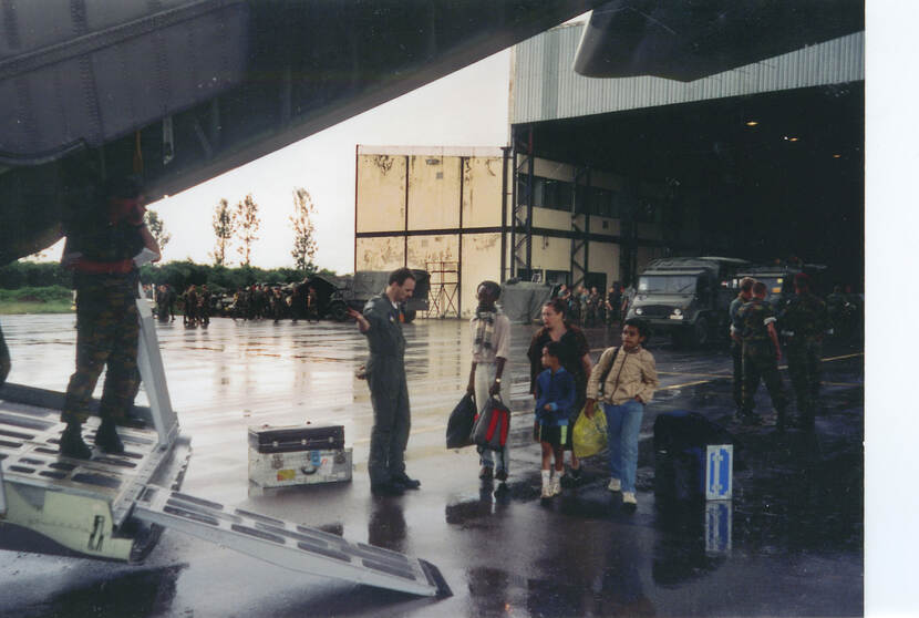 Kleurenfoto, een gevechtspiloot gebaart naar een man, een vrouw en twee kinderen om aan boord van een vliegtuig te stappen. Links op de loopplank van het vliegtuig staan twee militairen. Op de achtergrond een hangar, meerdere voertuigen en militairen.