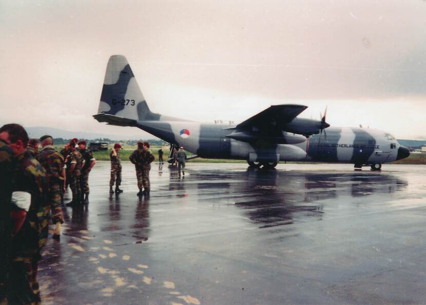 Kleurenfoto, een Nederlands transportvliegtuig van de Koninklijke Luchtmacht, in camouflagekleuren geschilderd, met de Nederlandse rondeel op het achterste deel. Links een groep militairen in camouflagekleding en rode baretten.