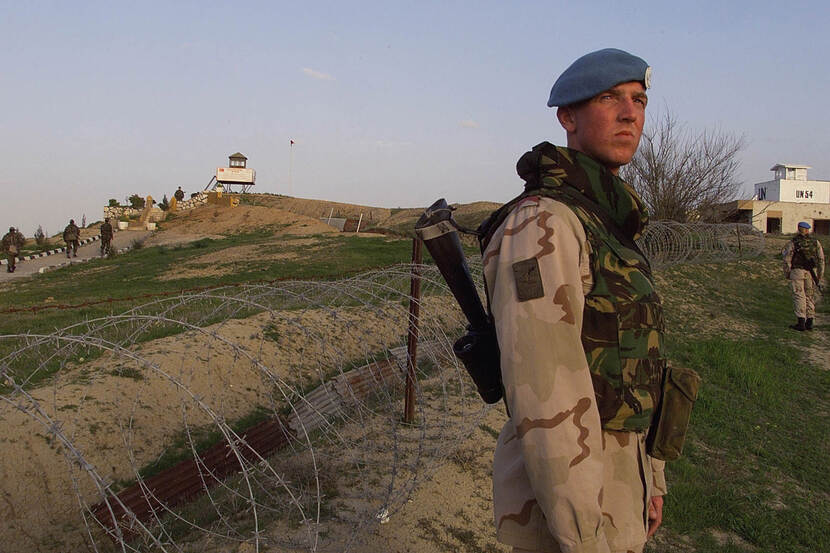 Kleurenfoto, een militair in camouflagekleding en een geweer kijkt over zijn rechterschouder richting de camera. Achter hem ligt prikkeldraad op de grond, die leidt tot een uitkijktoren op de top van de heuvel. Links op de achtergrond lopen drie mannen naar die uitkijktoren.