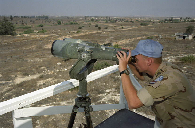Kleurenfoto, een militair in camouflagekleding en een lichtblauwe pet kijkt door een grote verrekijker. Hij staat achter een wit hekje. In de omgeving is een verdord landschap met struiken en enkele bomen te zien.