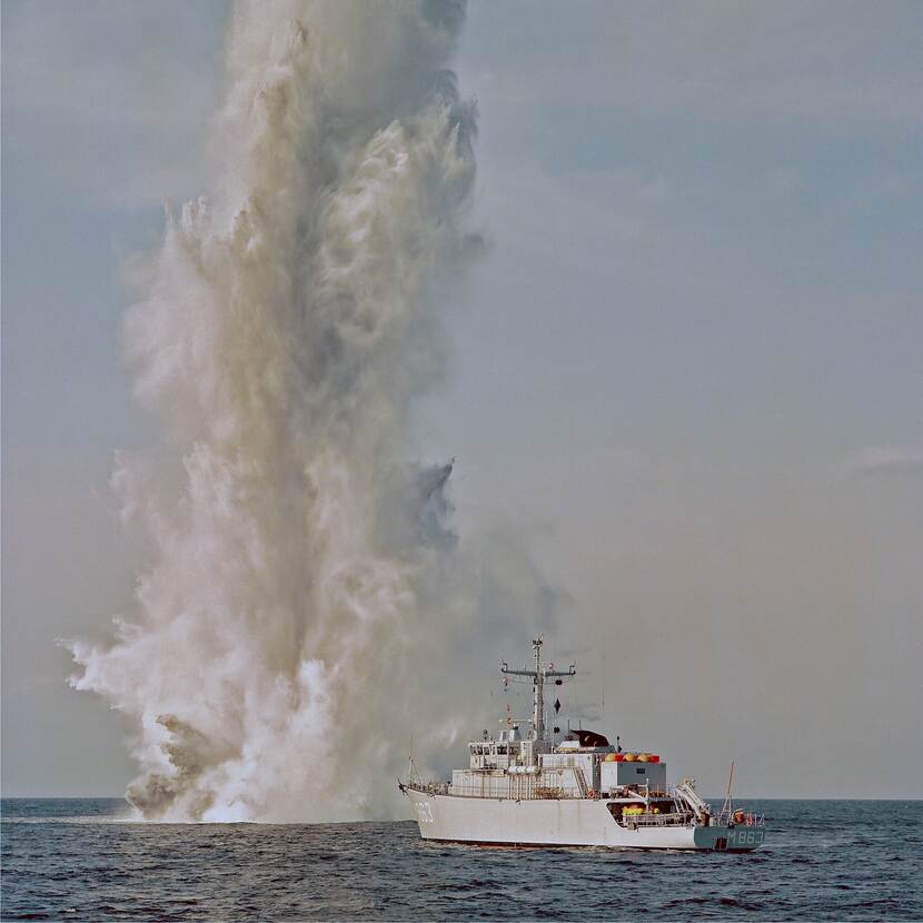Kleurenfoto, een marineschip op zee. Op de achtergrond een grote kolom water als gevolg van een onderzeese ontploffing.
