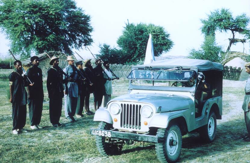 Kleurenfoto, een witte jeep met dak op de voorgrond, iemand zit achter het stuur en aan de zijkant is een stok met de VN-vlag vastgemaakt. Links een rij van acht militairen, waarvan zes een karabijn dragen. Op de achtergrond een hek, twee hutten en bomen.