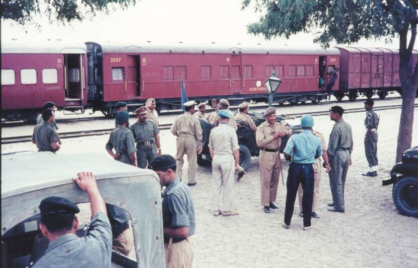Foto van een rode trein. Naast de trein staan meerdere militairen, waarvan één met blauwe helm en één met blauwe baret, en enkele jeeps.