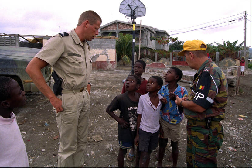 Kleurenfoto, buiten. Twee militairen, één in beige en de ander in camouflagekleding, praten op straat met vier kinderen. De linker militair draagt een holster met pistool aan zijn riem, de rechter heeft een gele pet op en op zijn mouw een zwarte lap met de letters MP in het wit. Op de achtergrond vervallen huizen en auto’s, een basket en lage bomen.