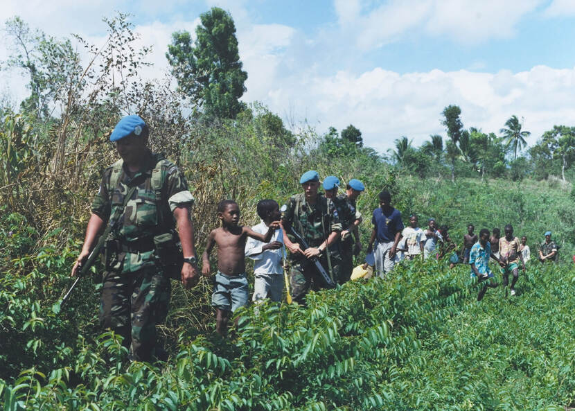 Kleurenfoto, buiten. Een rij van zestien personen loopt door struikgewas, voorop een militair in camouflagekleding, een blauwe baret en een geweer om zijn schouder. Daarachter twee kinderen, dan drie militairen (ook in camouflagekleding en met geweer) en meerdere mensen in burgerkleding. Om de rij heen veel struiken en bomen.