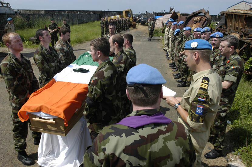 Kleurenfoto, buiten. Links in de voorgrond staan zes militairen die samen een doodskist dragen, over de doodkist is een vlag gedrapeerd, op de vla een baret. Rechts en links in de achtergrond staan militairen in een rij opgesteld. Rechts op de voorgrond leest een militair voor uit een boek. Verder in de achtergrond grote stukken verroest ijzer en een betonnen muur.