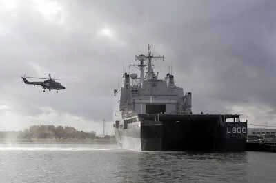 Kleurenfoto, buiten in een haven. Rechts ligt een groot marineschip in het water, van achteren bezien. Rechts op de achterkant staat L800 geschreven. Links van het schip hangt een helikopter in de lucht, onder het toestel stuift water omhoog. In de achtergrond bomen op de kade, de lucht is helemaal bewolkt.