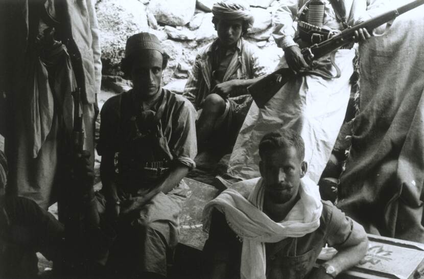 Zwart-witfoto, drie mannen zitten, twee staan, van deze twee is bovenste helft van hun lichaam buiten beeld, één van hen houdt een karabijn vast. Een Nederlandse man zit op de voorgrond en draagt een witte sjaal om zijn nek.