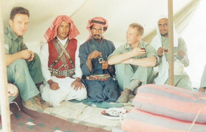 Kleurenfoto, vijf mannen zitten in een tent, dekens op de grond. Twee van hen dragen groene legerkleding, de andere drie lokale kleding, zoals tulbanden en versierde riemen. De twee mannen rechts houden een glaasje thee vast, de meest rechter man ook een theepot.