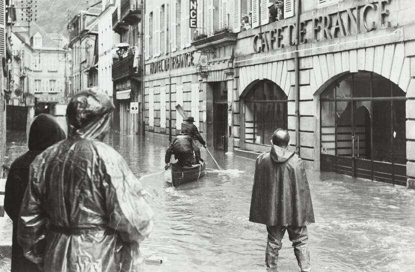 Zwart-witfoto, een ondergelopen straat, op het water varen twee mannen in een kano. 3 mannen in regenjassen staan op de voorgrond te kijken.