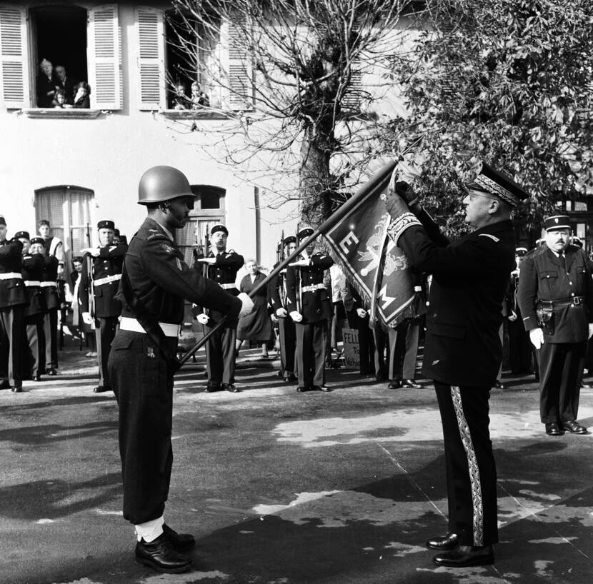 Zwart-witfoto, Franse militairen staan opgesteld in rijen. Op de voorgrond houdt 1 militair een vaandel vast terwijl de Prefect er een lint om hangt. Op de achtergrond kijken mensen naar de plechtigheid door hun ramen.