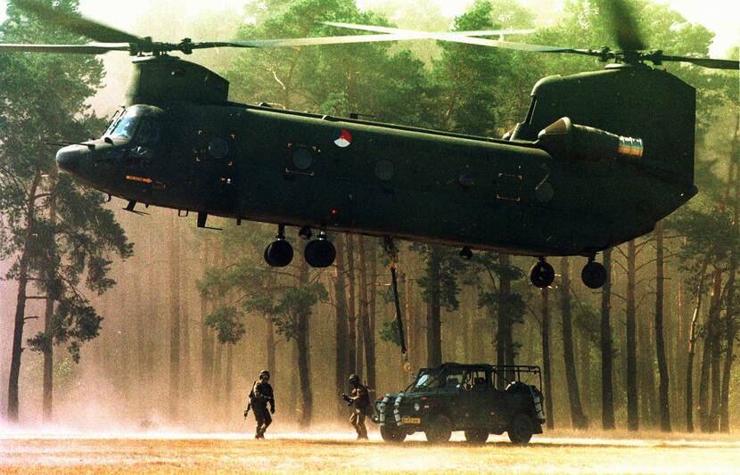 Kleurenfoto, een legergroene transporthelikopter hangt in de lucht, rotorbladen bovenop aan de voorkant en de achterkant. In het midden op de zijkant is de roundel van de Nederlandse luchtmacht geschilderd. Onder de helikopter op de grond staat een jeep, daarvoor twee militairen. Op de achtergrond bomen en opstuivend zand.
