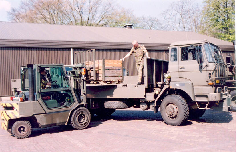 Kleurenfoto, rechts een vrachtwagen die wordt beladen, links een vorkheftruck die een houten kist op de laadbak van de vrachtwagen zet. In de laadbak staat een militair, achter het stuur van de vorkheftruck zit een andere militair. Op de achtergrond een lange hangar.