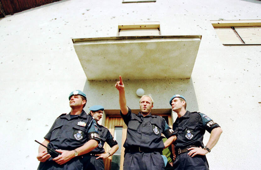 Kleurenfoto, van onderaf genomen. Vier mannen staan voor de ingang van een gebouw, boven hun hoofden een afdakje. Zij dragen donkerblauwe uniformen met om de linkermouw een band met het woord Police erop, drie hebben een baret op. De man zonder baret wijst met zijn rechterarm naar voren, de man links heeft een portofoon vast.