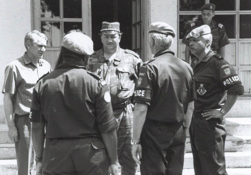 Zwart-witfoto. Een groep van vijf mannen staat buiten voor de ingang van een gebouw te praten. Drie dragen militaire uniformen, inclusief een baret, en een band om de linkermouw waar Police op staat. De middelste persoon draagt een ander militair uniform. Links een man in burgerkleding. Op de achtergrond staat nog een militair op de trap naar de deur.