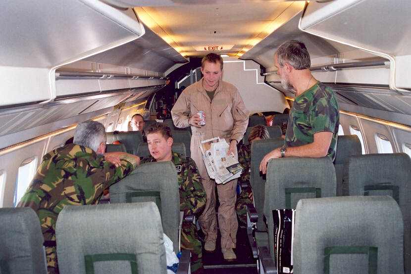 Kleurenfoto, binnen in een vliegtuig. Zes militairen zitten en staan in een vliegtuig. In het midden een militair in beige overall, met in zijn ene hand een blikje cola en in de andere een stapeltje kranten. Alle andere militairen dragen camouflagekleding. Rechts staat een militair tussen de stoelen, hij kijkt naar de militair in het gangpad. Links zit een militair achterstevoren en leunt op de rugleuningen voor hem, hij praat met een militair op de rij daarachter.