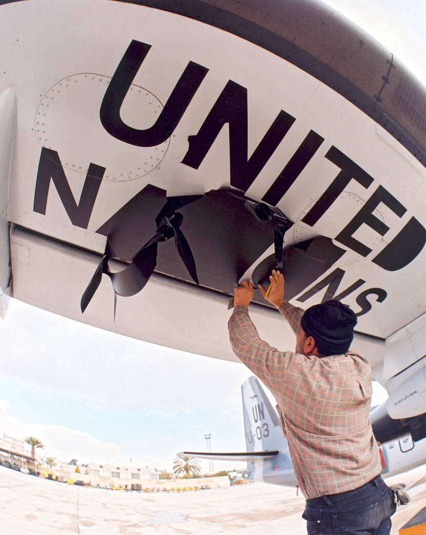 Kleurenfoto. Een man met een muts staat onder een vliegtuigvleugel. Boven zijn hoofd staat in zwarte letters het woord ‘United’. Hij snijdt in een zwart vlak van plastic, links daarvan de letter N, rechts de letters NS. In de achtergrond de staart van het vliegtuig, palmbomen en gebouwen van een vliegveld.