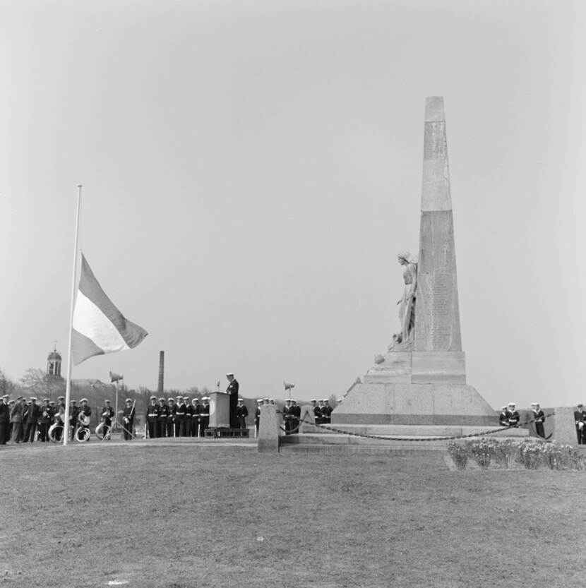 Zwart-wit foto. Officieren en manschappen van de vloot en Korps Mariniers herdenken de gevallenen van de marine en de koopvaardij tijdens Dodenherdenking bij het oorlogsmonument 'Voor hen die vielen' op het Havenplein te Den Helder. Verder zijn aangetreden muzikanten van de Marinierskapel en de gewapende wacht. Toespraak wordt gehouden door de vlootaalmoezenier.