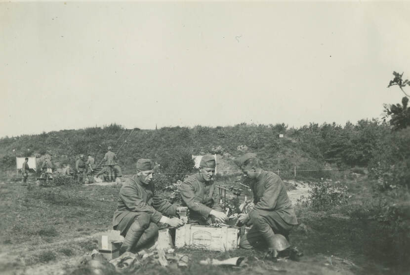 Zwart-witfoto. Drie Nederlandse militairen zitten op de grond met een houten kistje tussen hen in, op de grond ligt rommel, de linker houdt een stapeltje blokjes overeind, de andere twee vullen een rond mitrailleurmagazijn. Links in de achtergrond andere militairen met een kanon, tegen de begroeide dijk daarachter zijn witte doelwitten aangebracht.