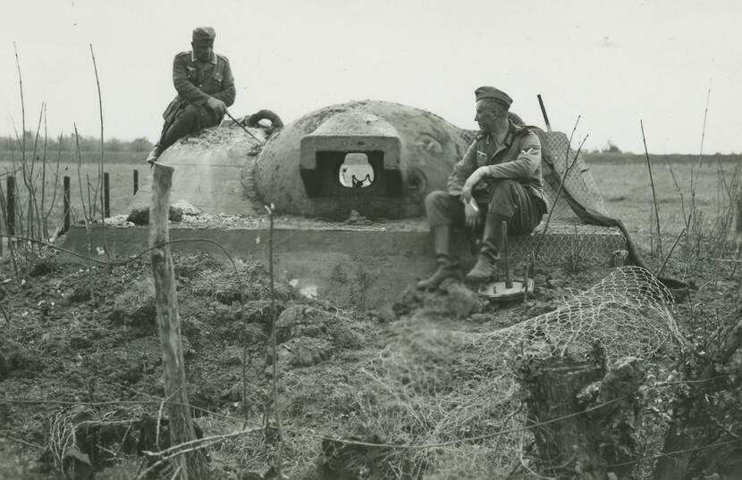 Zwart-witfoto. Twee Duitse militairen zitten bovenop een bunker en kijken naar de betonnen koepel, de linker wijst met een stok naar de koepel. Om hen heen beschadigd hekwerk, palen en veel aarde, in de achtergrond weilanden.