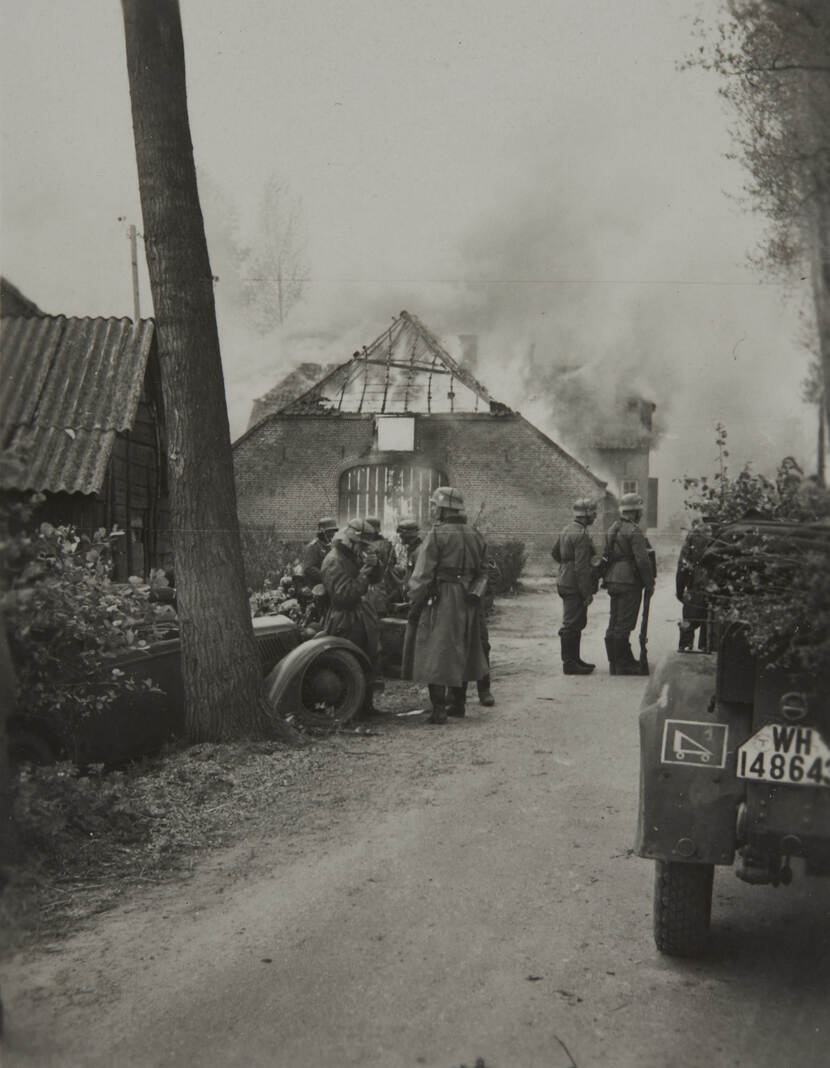 Zwart-witfoto. Een boerderij staat in brand, de vlammen schieten door het geraamte van het dak en de poort. Daarvoor staan zeven Duitse militairen, om hen heen met bladeren en takken bedekte auto’s en motoren.