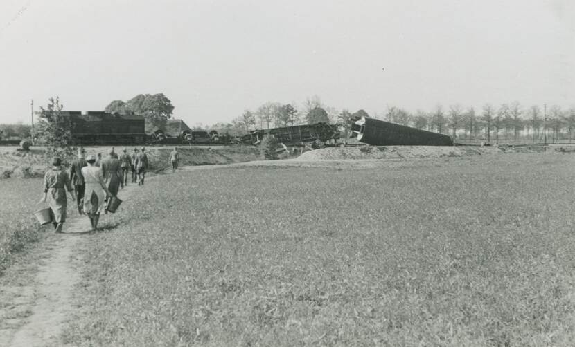 Zwart-witfoto. Polderlandschap met een verhoogde spoorbaan, daarop staat een trein, drie wagons zijn ontspoord en liggen op hun kant. Links loopt een groep van tien mensen naar de wrakstukken, de achterste twee zijn vrouwen.