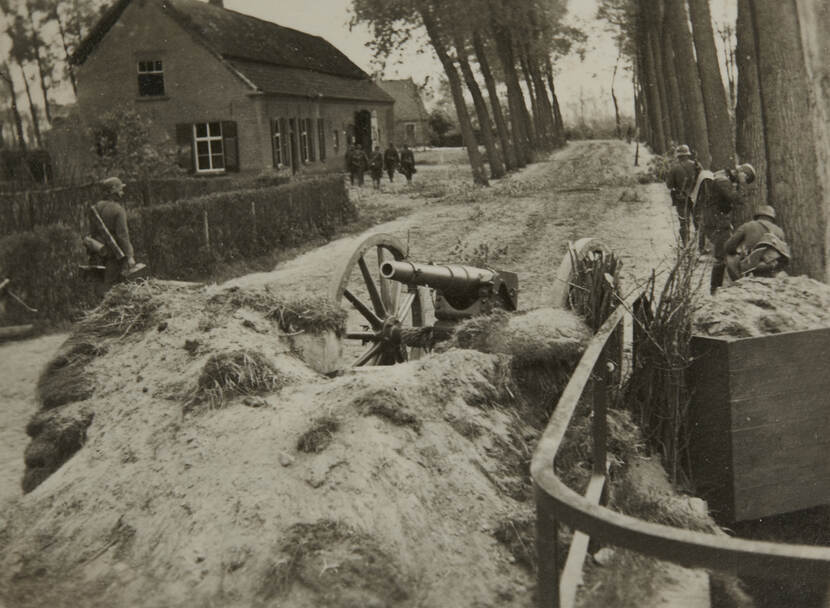 Zwart-witfoto. Een kanon staat verlaten achter een aarden stelling aan een onverharde weg, rechts staan drie Duitse militairen lag de bomenrij, links loopt nog een Duitse militair. In de achtergrond lopen vijf militairen naar de boerderij langs de weg.