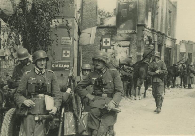 Zwart-witfoto. Straatbeeld, links op de voorgrond leunen vier Duitse militairen op een stuk geschut, rechts in de achtergrond enkele Duitse militairen te paard. De gebouwen in de achtergrond zijn beschadigd.