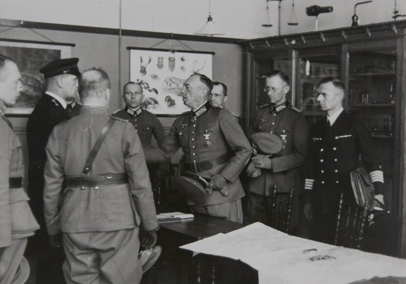 Zwart-witfoto. Acht militairen staan in een klaslokaal, allemaal in vol ceremonieel uniform gekleed, op één na houdt iedereen zijn pet in de hand. De vierde van rechts schud de hand van een militair die links half verscholen achter een andere staat. Tegen de muren boekenkast en lesplaten, tussen de militairen een tafel met stoelen ernaast en een kaart erop.