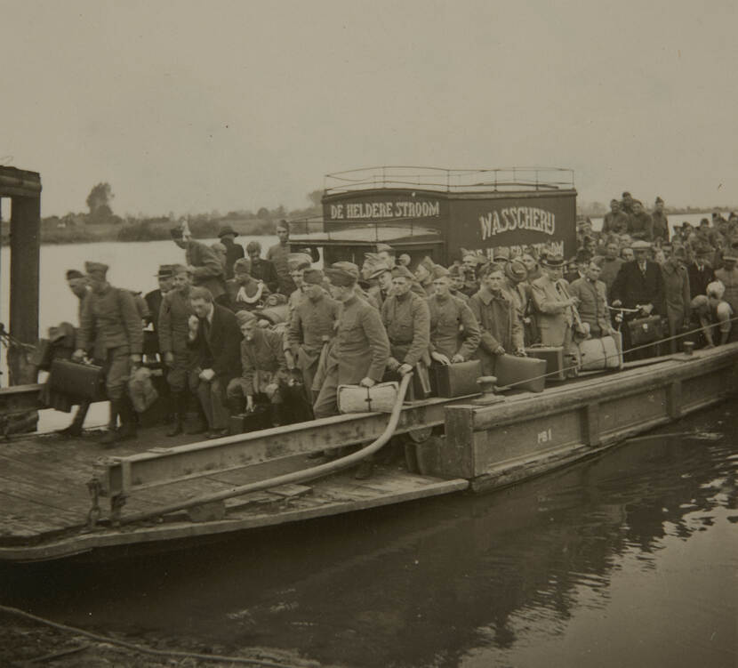 Zwart-witfoto. Een veerboot staat op het punt van aanmeren, aan boord zijn tientallen mensen op elkaar gepakt, velen van hen dragen een militair uniform, bijna iedereen heeft een koffer bij zich. Tussen de mensen staat een vrachtwagen van een wasserette.